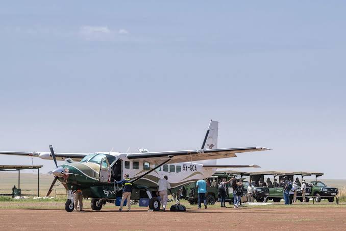 A Cessna Caravan 208 on a dirt bush airstrip with passengers, bags and safari vehicles